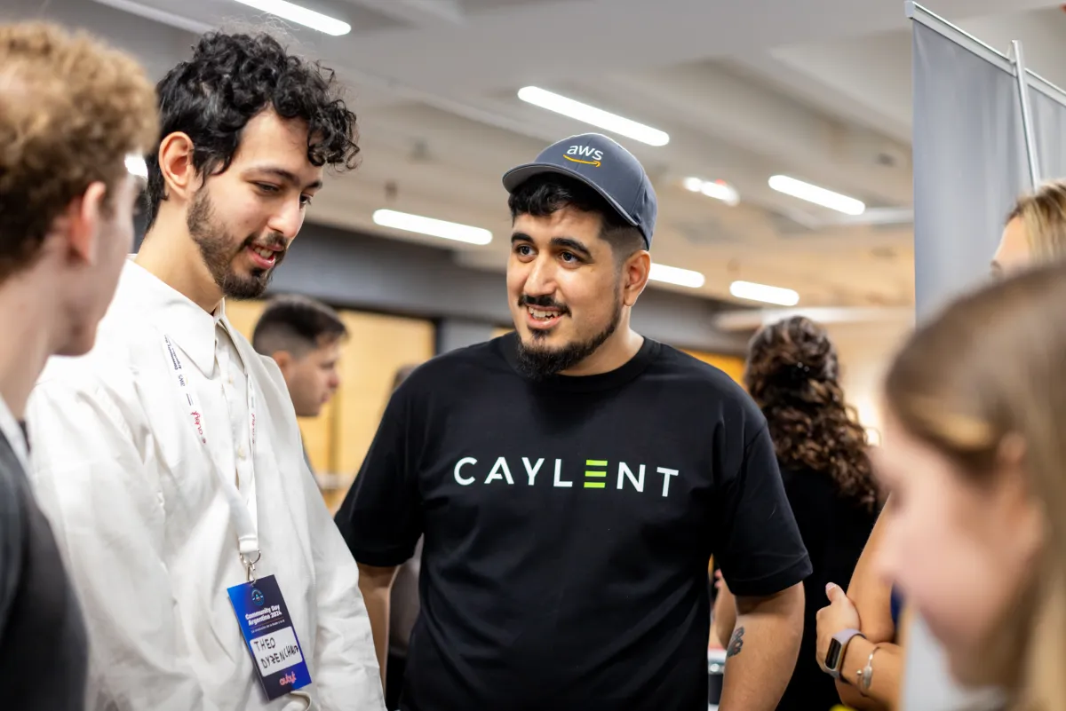Asistentes haciendo networking, uno con camiseta Caylent y gorra AWS